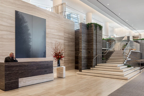 A modern lobby with a marble reception desk, a person on the phone, a large minimalist painting, floral arrangement, and a grand staircase with glass railings. The space is bright with neutral tones and sleek design.