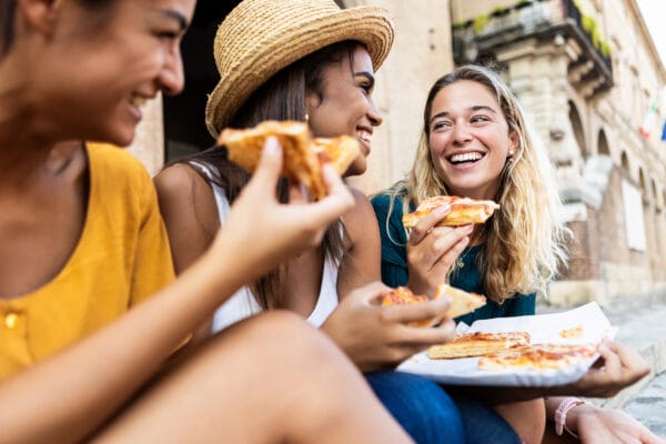 Three young multiracial women laughing while eating a piece of pizza in Italy
