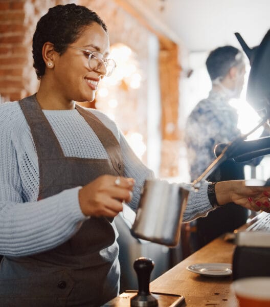 Beautiful Latin American Female Barista with Short Hair and Glasses is Making a Cup of Tasty Cappuccino in Coffee Shop Bar. Portrait of Happy Employee Behind Cozy Loft-Style Cafe Counter.