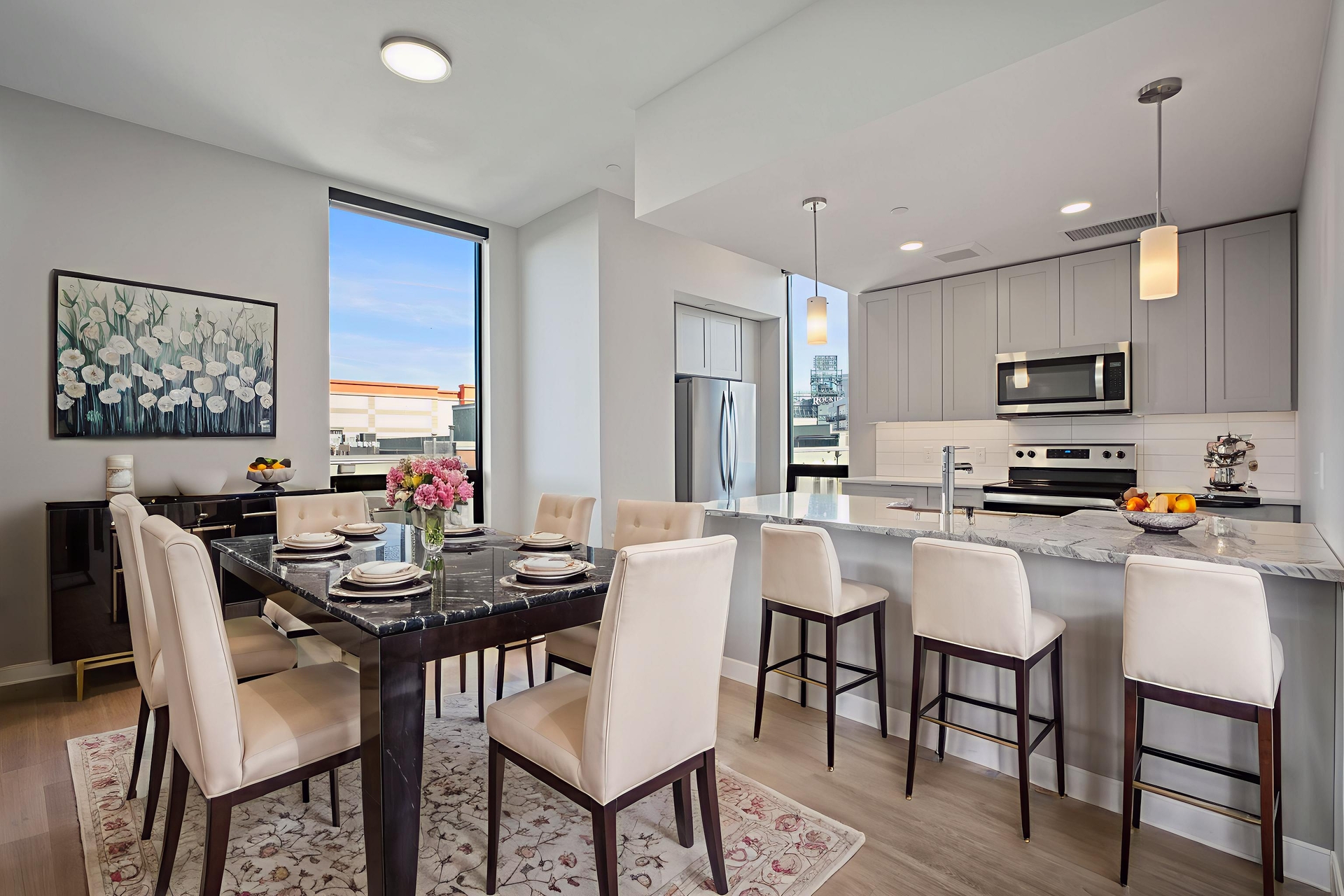 Modern kitchen and dining area at Windsor 3000 Huron with light gray cabinets, stainless steel appliances, marble countertops, a dark wood dining table for six, white cushioned chairs, large windows, and floral artwork on the wall.