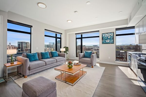 A modern, bright living room at Windsor 3000 Huron with large windows overlooking a cityscape, featuring a gray sofa, blue cushions, armchair, wooden coffee table, and contemporary decor. Natural light fills the space.